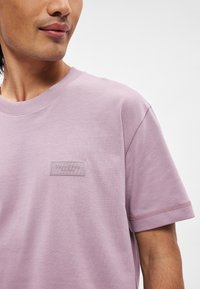 T-shirt di cotone lilla con scollo rotondo, maniche corte e un'etichetta di marca texturizzata sul petto. Finitura del tessuto semplice e liscia.