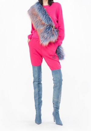 Femme portant un sweat-shirt et un short rose vif, des cuissardes en daim bleu et une large écharpe en fausse fourrure bleue et orange traversant sa poitrine.