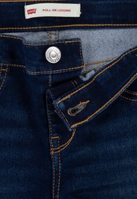 Leggings in denim blu scuro con cintura elasticizzata, caratterizzati da un bottone a pressione argentato, cuciture dorate a contrasto e un'etichetta in vita. Texture liscia.