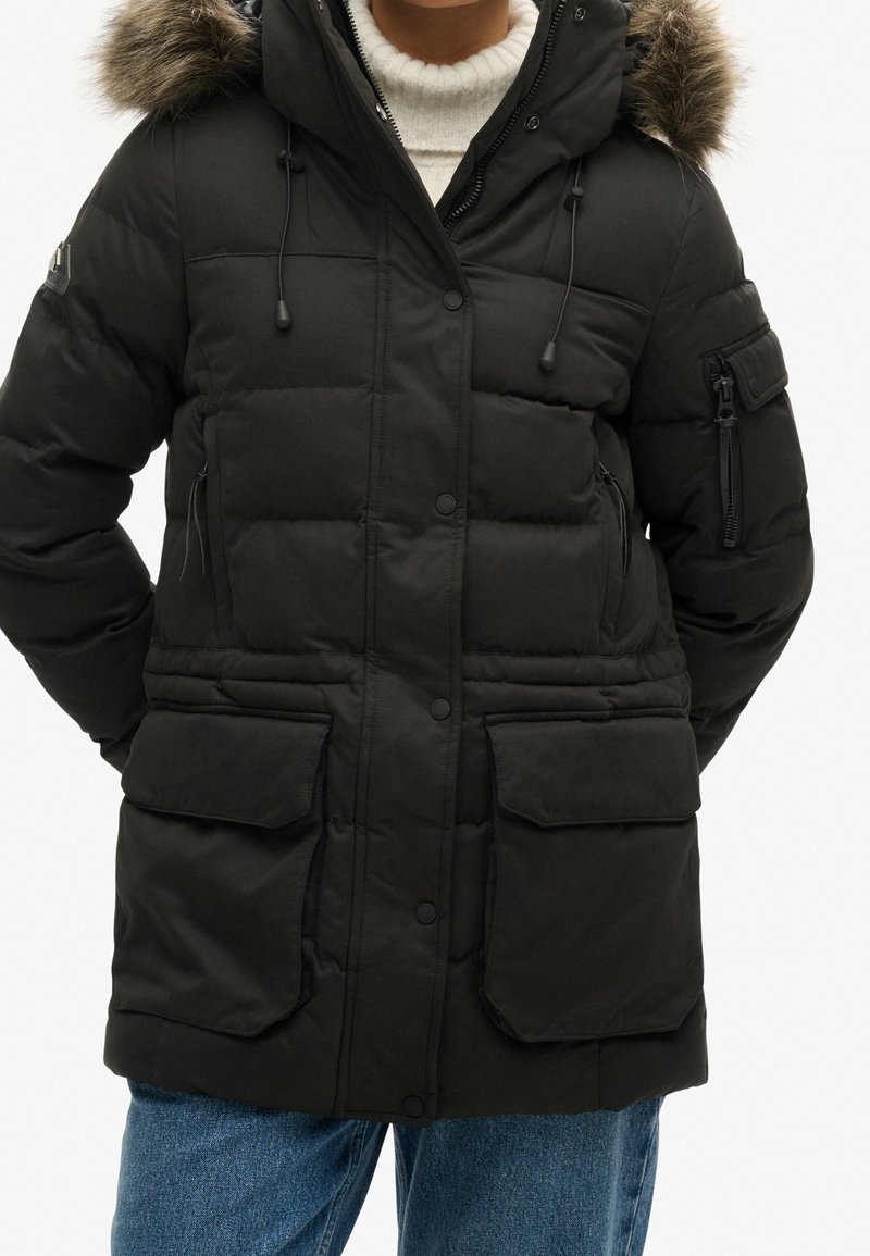 Persona con un abrigo de invierno acolchado negro con capucha forrada de piel, bolsillos con solapa y bolsillo con cremallera en el brazo, combinados con jeans azules y un suéter blanco.