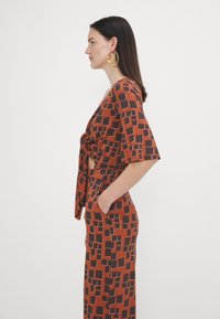 Hnědý vzorovaný dvoudílný outfit s krátkými širokými rukávy. Obsahuje uzlový přední díl a boční kapsy; hladká textura s geometrickými tvary.