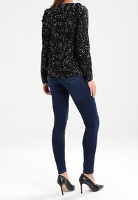 Blouse noire avec un motif à pois blancs, détail volanté aux épaules, associée à un jean slim bleu foncé et des chaussures à talons noirs.