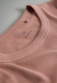 Tricou roz cu dungi, cu decolteu rotund, având cusături vizibile și o etichetă pe care scrie „THE SET. Fabricat în Bangladesh.” Textură moale.