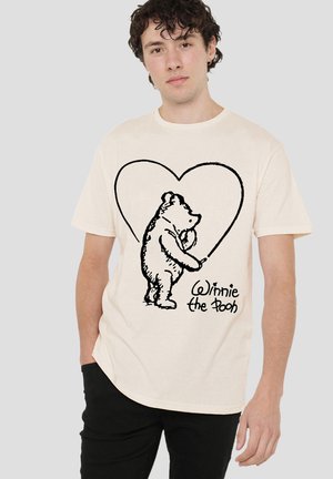 Junger Mann trägt beige T-Shirt mit schwarzer Skizze von Winnie Puuh, der ein Herz zeichnet und dem Text „Winnie Puuh“.