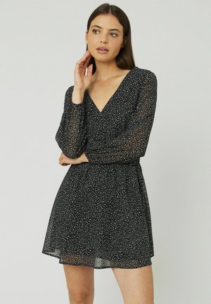 Robe noire avec de longues manches transparentes et un décolleté en V, ornée d'un motif à pois blancs et d'une taille froncée pour une silhouette évasée.