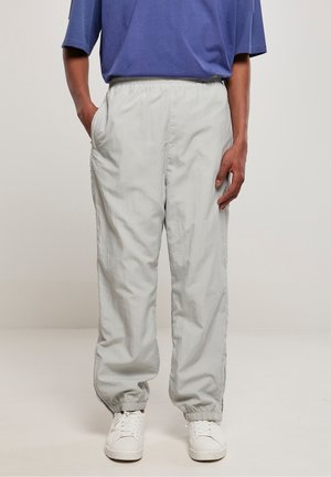 Personne portant un pantalon gris clair à taille élastique et poignets élastiques, des baskets blanches et une chemise bleue, debout avec une main dans la poche.