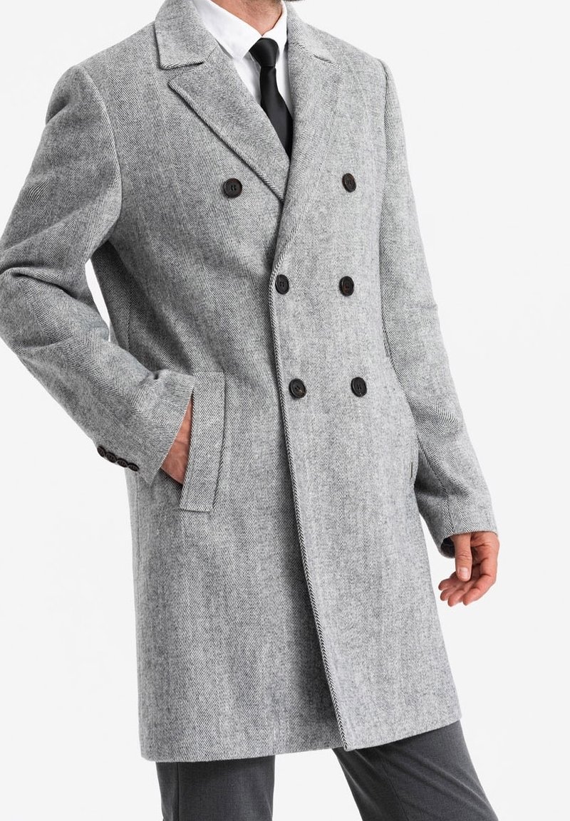 Homme portant un manteau gris en laine à double boutonnage avec des boutons noirs, une chemise blanche, une cravate noire et un pantalon gris, main dans la poche.