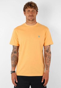 T-shirt à manches courtes, col rond en coton orange clair. Présente un petit logo bleu sur la poitrine. Texture lisse, coupe décontractée, design basique.