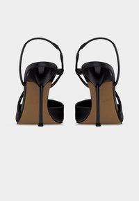 Ζευγάρι μαύρα γόβες με λουράκι στο πίσω μέρος, εμφανιζόμενα από πίσω σε απλό λευκό φόντο.