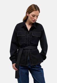 Chemise noire avec un col, deux poches poitrine et une taille ceinturée. Présente des coutures blanches contrastantes et des manches longues. Pantalon en denim.