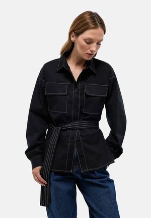 Chemise noire avec un col, deux poches poitrine et une taille ceinturée. Présente des coutures blanches contrastantes et des manches longues. Pantalon en denim.