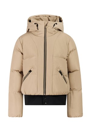 Kind draagt een beige donsjack en broek, zwarte handschoenen, zwarte muts en zwarte laarzen, staat en lacht op een lichte achtergrond.