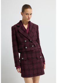 Blazer en tweed bordeaux au design court, devant croisé et boutons argentés. Associé à une jupe à carreaux assortie.