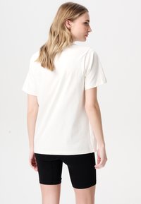 Crème-kleurig korte mouwen t-shirt in een ontspannen ontwerp, gedragen met zwarte geribbelde fietsshorts, gezien vanuit de achterkant.
