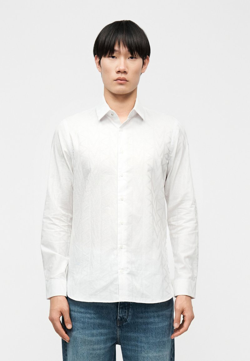 Νεαρός άνδρας με ίσια μαύρα μαλλιά φοράει λευκό πουκάμισο με κουμπιά και μπλε τζιν, στέκεται μπροστά σε απλό ανοιχτό φόντο.