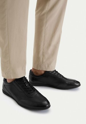 Černé nízké kožené tenisky s perforovanými detaily, nošené s béžovými kalhotami na světlém pozadí.