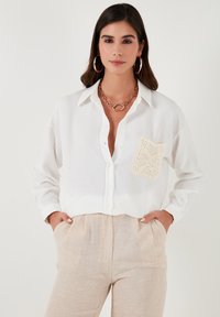 Camisa blanca de botones y mangas largas, con un bolsillo de crochet en crema texturizado. Combinada con pantalones texturizados en un tono beige claro.