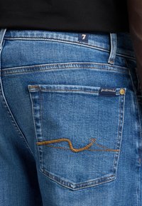 Niebieskie dżinsy z tylną kieszenią, która ma kontrastujące brązowe zygzakowate przeszycie oraz małą metkę z logo marki. Tkanina ma wyblakłą teksturę.