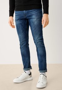 Hombre con vaqueros azules remangados, calcetines blancos y zapatillas blancas de pie sobre un suelo beige con un fondo claro.