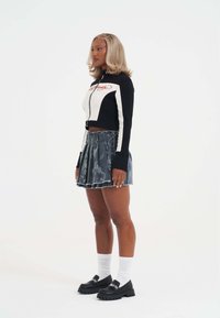 Ung kvinde med blond hår iført en sort og hvid cropped jakke, plisseret denim miniskørt, hvide sokker og sorte chunky loafers, står sidelæns.