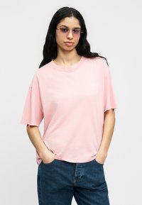 Γυναίκα με μακριά σκούρα μαλλιά που φορά ροζ γυαλιά ηλίου με ροζ απόχρωση, χαλαρή ανοιχτό ροζ μπλούζα και μπλε τζιν, στέκεται με τα χέρια στις τσέπες.