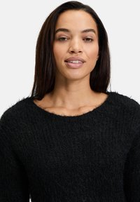 Schwarzer gestrickter Pullover mit lockerer Passform, Rundhalsausschnitt und einer weichen, flauschigen Textur. Der Stoff hat ein geripptes Muster und leichte Fransen am Ausschnitt.