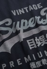 Camiseta de algodón negra con texto en estilo vintage en color gris, incluyendo la palabra "Super" y otros caracteres, con un acabado suave y ligeramente texturizado.
