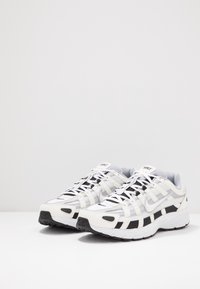 Sportovní boty s bílým syntetickým svrškem, černými akcenty, texturovanou síťovinou a gumovou podešví, s krajkovým designem a polstrovaným límcem.
