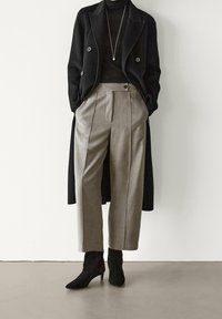 Manteau noir en laine à double boutonnage, col roulé noir ajusté, pantalon gris à jambes larges avec plis, et bottines en daim noir.