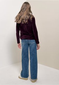 Frau mit langen Haaren, die ein dunkelpurpurnes langärmliges Oberteil, breite blaue Jeans und beige Sneakers trägt, steht vor einer beigen Wand.