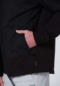 Chaqueta negra con un bolsillo con cremallera, dobladillo ajustable y puños elásticos. El material tiene una textura suave con un acabado mate.