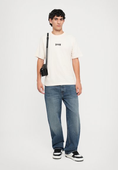 T-shirt crema con grafica 'BARROW', abbinata a jeans in denim blu larghi e sneaker bianco e nero, indossata con una piccola borsa nera.
