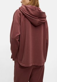 Oversized maroon hoodie with a hood, smooth texture, casual design, and dropped shoulders, featured on a model from a rear angle.