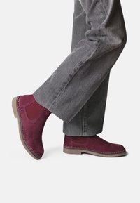 Burgundy mockaskstövlar med elastiska sidopaneler, sydda detaljer och en gummisula, kombinerade med grå vida byxor.