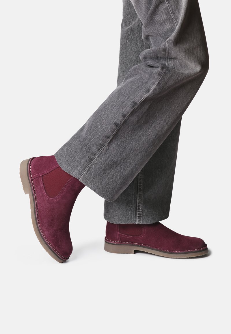 Burgundy mockaskstövlar med elastiska sidopaneler, sydda detaljer och en gummisula, kombinerade med grå vida byxor.