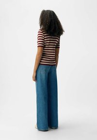 Haut ajusté rayé bordeaux et beige associé à un jean en denim bleu ample à jambes larges. Cheveux bouclés visibles de dos, posé contre un fond uni.