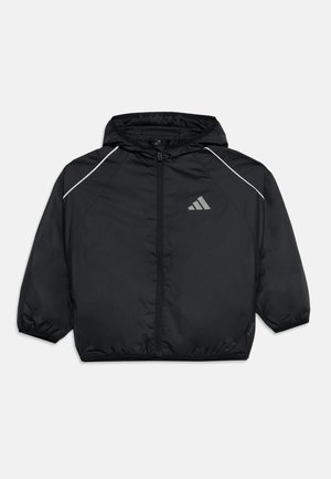 Veste noire à capuche avec fermeture éclair devant, poignets élastiques, passepoil blanc aux épaules et petit logo réfléchissant sur la poitrine.
