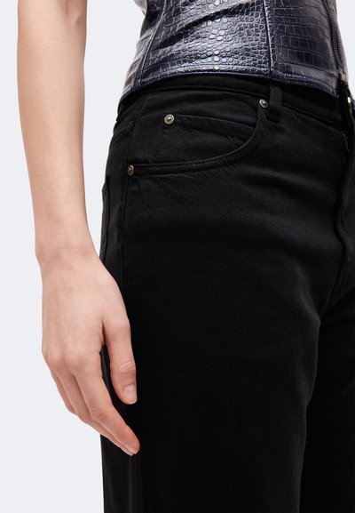 Gros plan sur une personne portant un jean noir et une ceinture noire texturée, une main posée à côté de la cuisse.