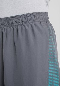 Detailní záběr na šedé elastické šortky v pase s tyrkysovým tečkovaným vzorem po stranách, nošené s světle šedým tričkem.