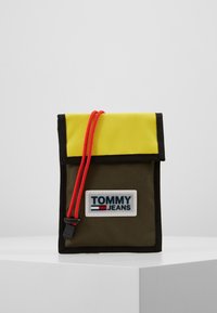 Borsa in tessuto verde con un flap giallo, cordino rosso e accenti neri. Presenta un'impronta del logo "TOMMY JEANS" sul davanti.