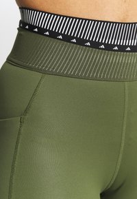 Zelené sportovní legíny mají široký pás s texturou v černobílém pruhu a malými trojúhelníkovými akcenty a bočními kapsami.