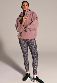 Giacca leggera mauve con cappuccio e polsini elasticizzati abbinata a leggings neri con un motivo astratto viola e scarpe da ginnastica nere.