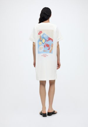 Kobieta z długimi ciemnymi włosami, ubrana w białą sukienkę w stylu T-shirt z kolorową ilustracją stołu na imprezę z rakami na plecach.