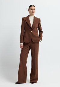 Completo marrone su misura caratterizzato da un blazer strutturato con un singolo bottone dorato, abbinato a pantaloni a gamba larga. Tessuto liscio e design elegante.