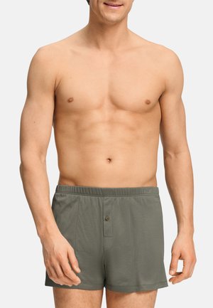 Uomo senza maglietta che indossa boxer grigi con vita elastica e patta con un solo bottone, in piedi davanti a uno sfondo chiaro uniforme.
