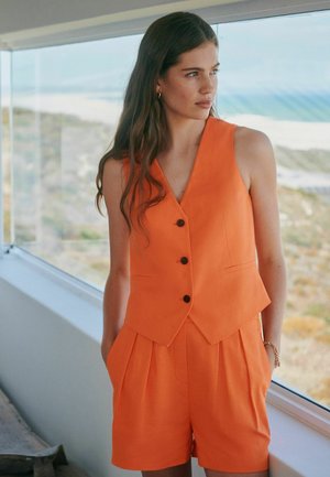 Woman with long hair wearing an orange sleeveless buttoned vest and matching high-waisted shorts, standing by a window overlooking a coastal landscape.