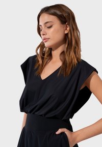 Femme aux cheveux bruns mi-longs portant une robe noire sans manches à encolure en V avec une taille cintrée, regardant vers le bas, main dans la poche.