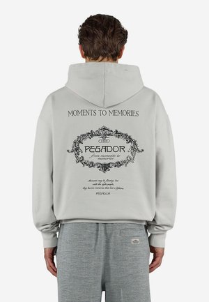 Mand iført lyst grå hættetrøje med sort udsmykket tekstdesign på ryggen, der siger "Moments to Memories" og "PEGADOR" med dekorativ ramme.
