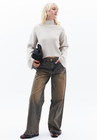 Beige Ripppullover mit weiten Ärmeln, kombiniert mit ausgewaschenen, locker sitzenden blauen Jeans mit fransigem Saum und einer schwarzen Leder-Clutch.