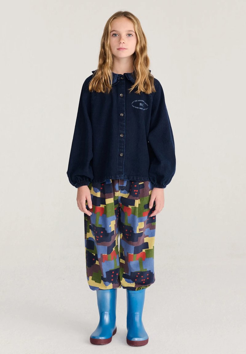 Jeune personne aux longs cheveux blonds se tient debout, portant une chemise sombre boutonnée, un pantalon coloré à motifs et des bottes en caoutchouc bleu vif.